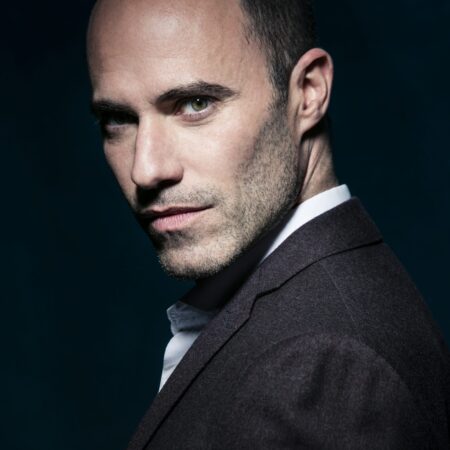 Leigh Melrose, a man with short hair and light stubble, wearing a dark suit jacket over a white shirt, looks confidently over his shoulder against a dark, blue-toned background.