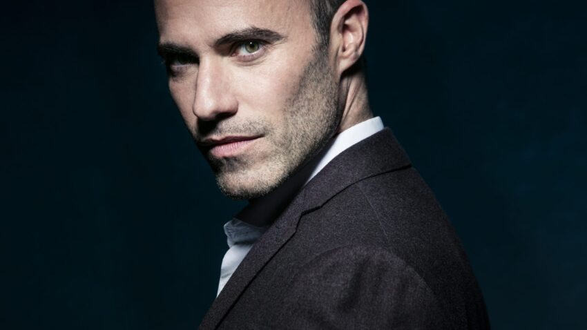 Leigh Melrose, a man with short hair and light stubble, wearing a dark suit jacket over a white shirt, looks confidently over his shoulder against a dark, blue-toned background.
