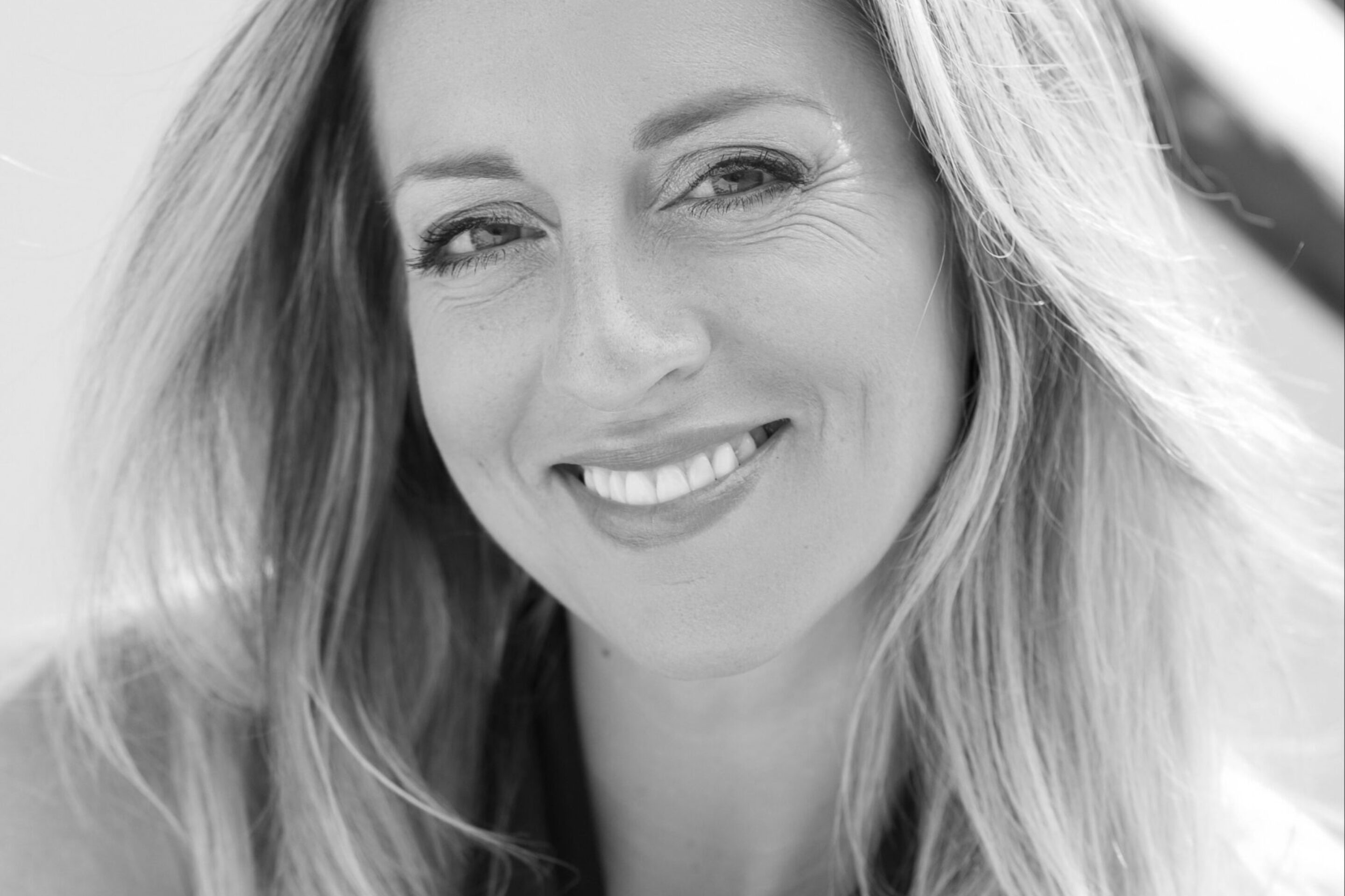 A woman with long, fair hair smiles warmly at the camera. She is wearing a sleeveless, dark top, and the photo is in black and white. The background is softly blurred.