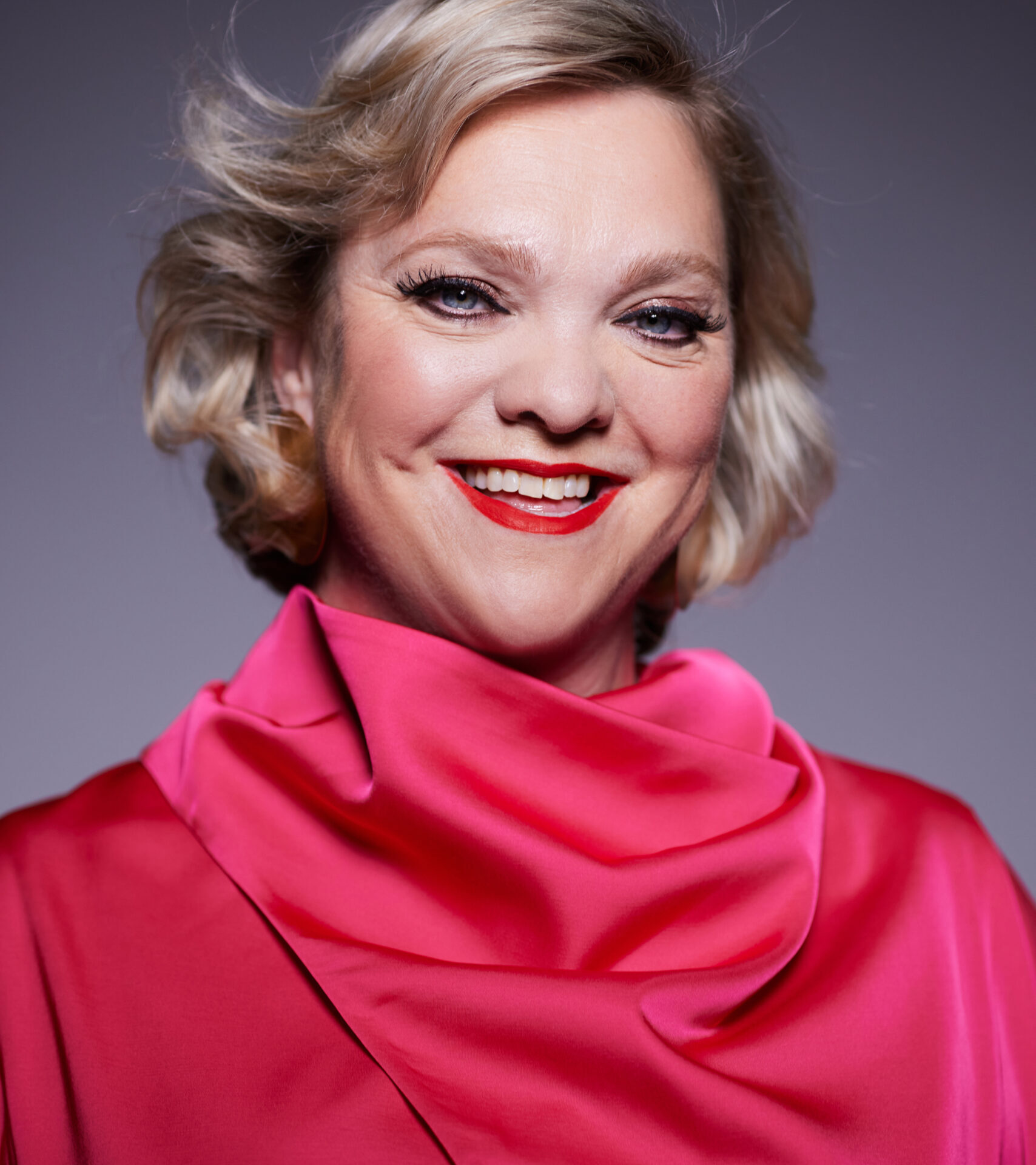 A smiling person with short blonde hair is wearing a bright pink, high-collared blouse. The softly lit background highlights both their cheerful expression and vibrant style, reminiscent of Madeleine Shaw’s uplifting presence.