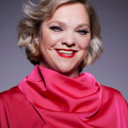 A smiling person with short blonde hair is wearing a bright pink, high-collared blouse. The softly lit background highlights both their cheerful expression and vibrant style, reminiscent of Madeleine Shaw’s uplifting presence.
