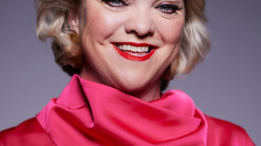 A smiling person with short blonde hair is wearing a bright pink, high-collared blouse. The softly lit background highlights both their cheerful expression and vibrant style, reminiscent of Madeleine Shaw’s uplifting presence.