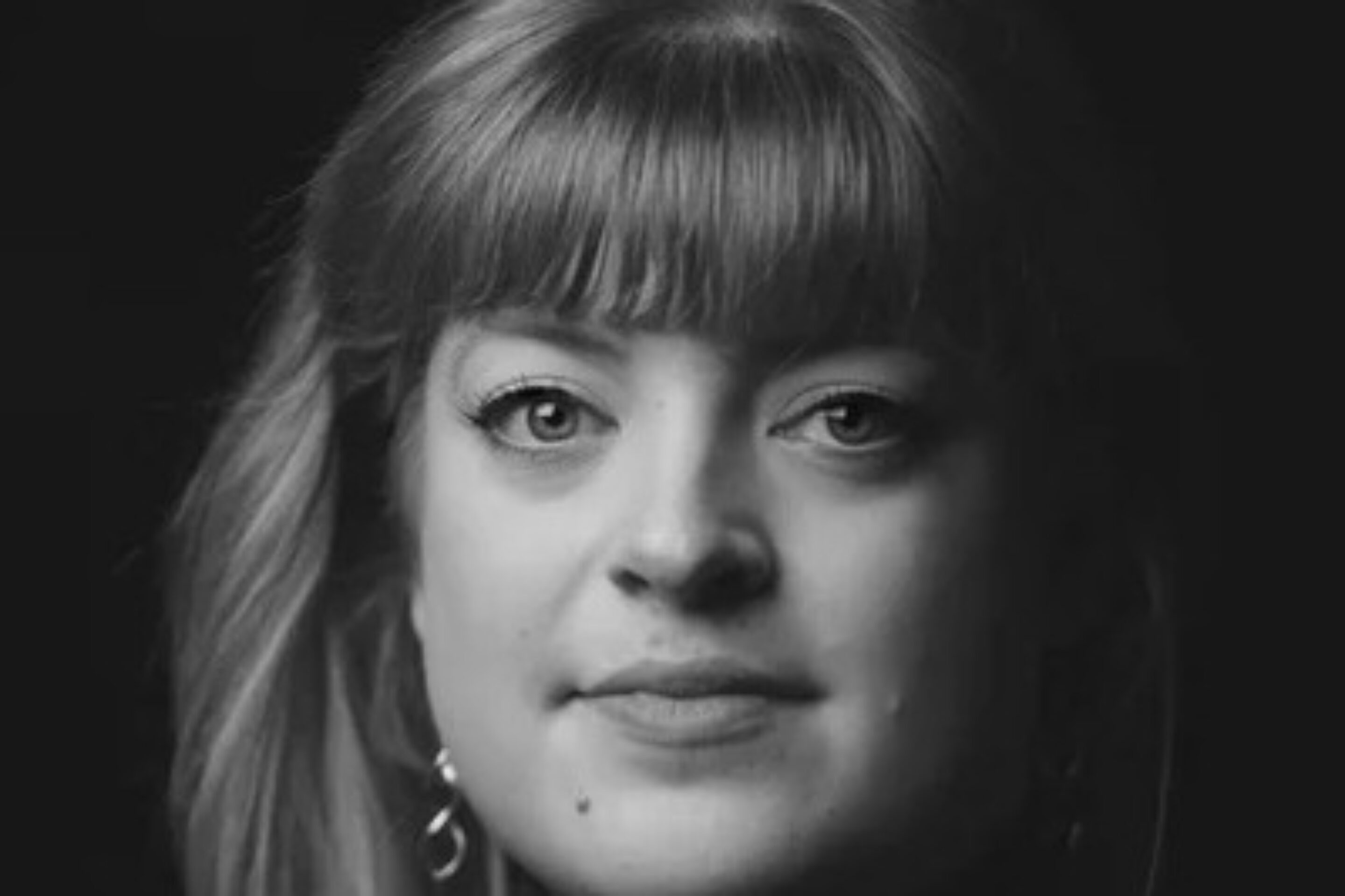 Black and white portrait of Madeline Boreham, a woman with fair hair and fringe, wearing earrings and a dark polo neck, looking directly at the camera with a neutral expression against a dark background.