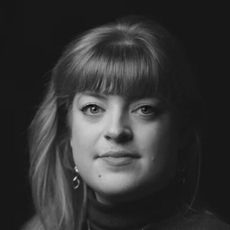 Black and white portrait of Madeline Boreham, a woman with fair hair and fringe, wearing earrings and a dark polo neck, looking directly at the camera with a neutral expression against a dark background.