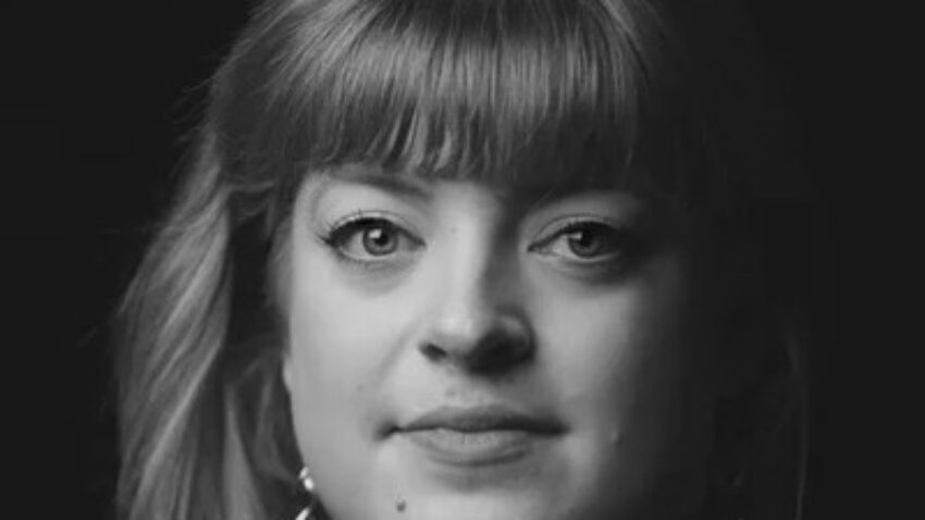 Black and white portrait of Madeline Boreham, a woman with fair hair and fringe, wearing earrings and a dark polo neck, looking directly at the camera with a neutral expression against a dark background.