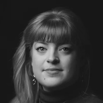 Black and white portrait of Madeline Boreham, a woman with fair hair and fringe, wearing earrings and a dark polo neck, looking directly at the camera with a neutral expression against a dark background.