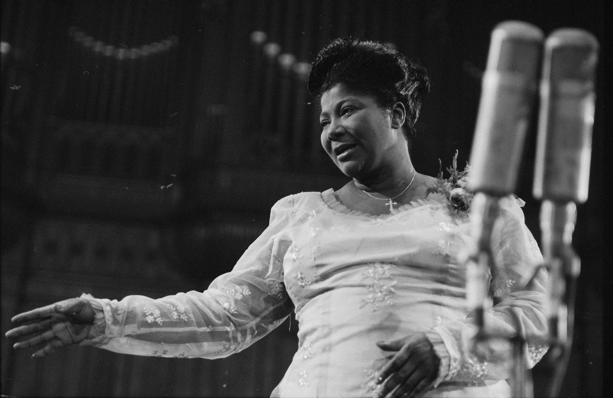 A woman in a light, embroidered dress sings "Summertime" by George Gershwin passionately on stage, standing near two microphones with one arm extended. The dark background highlights her and the microphones.