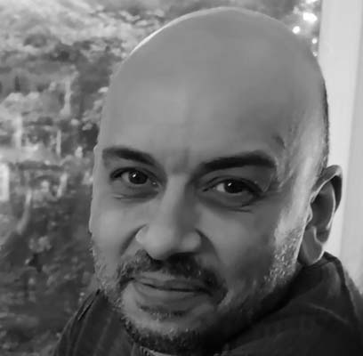 Marc Rosette, a man with a shaven head and trimmed beard, is looking at the camera and smiling softly. He is indoors near a window, with blurred greenery visible outside. The black and white photo captures his calm presence.