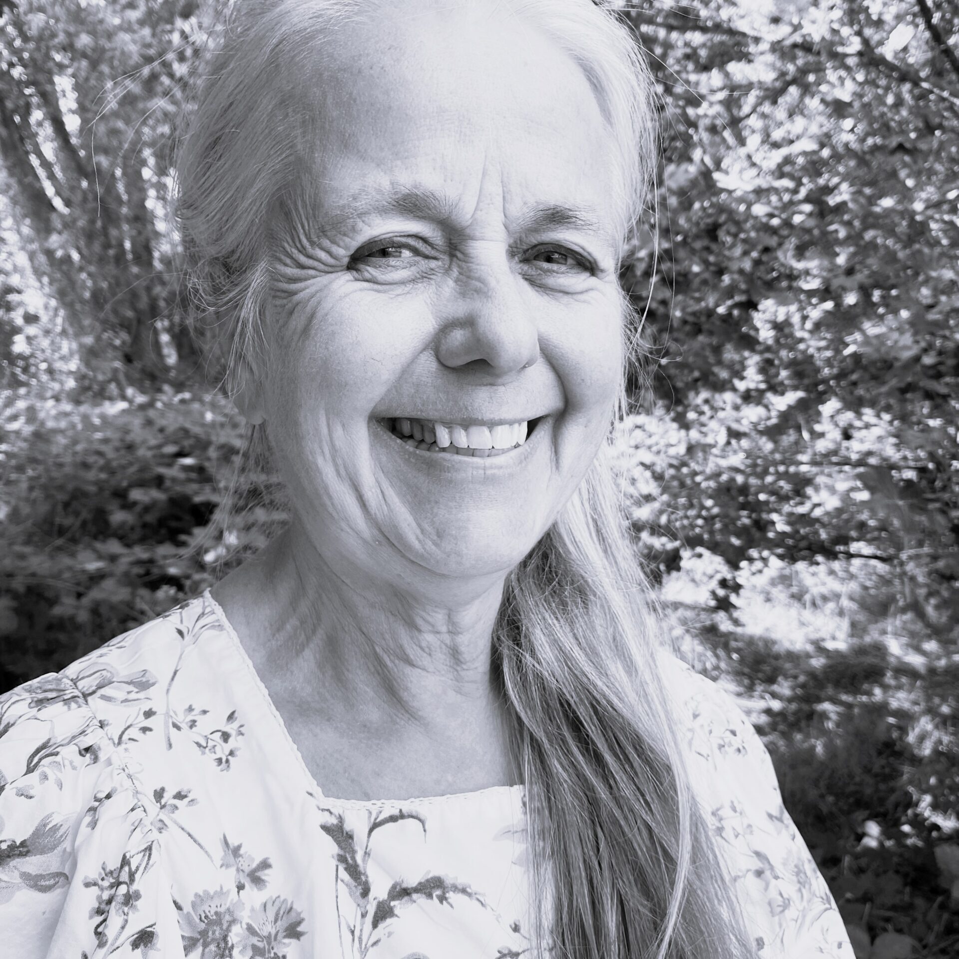 Margaret Roseberry, a smiling older woman with long grey hair in a floral blouse, stands outdoors surrounded by leafy trees and natural light. The photo is in black and white.