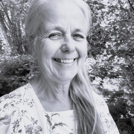 Margaret Roseberry, a smiling older woman with long grey hair in a floral blouse, stands outdoors surrounded by leafy trees and natural light. The photo is in black and white.