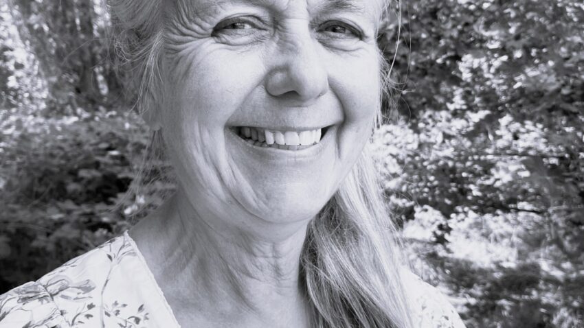 Margaret Roseberry, a smiling older woman with long grey hair in a floral blouse, stands outdoors surrounded by leafy trees and natural light. The photo is in black and white.