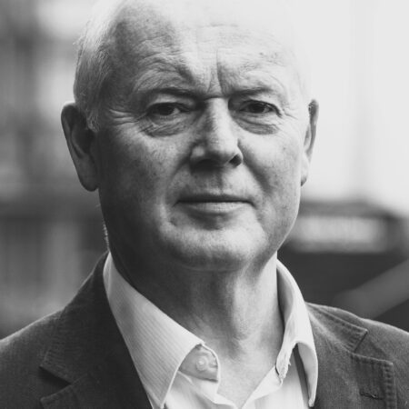 A black and white portrait of Mark Le Brocq, an older man with short, fair hair, wearing a collared shirt and blazer, looking directly at the camera with a neutral expression.