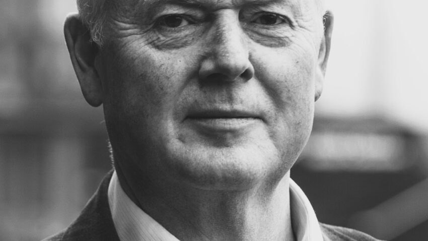 A black and white portrait of Mark Le Brocq, an older man with short, fair hair, wearing a collared shirt and blazer, looking directly at the camera with a neutral expression.