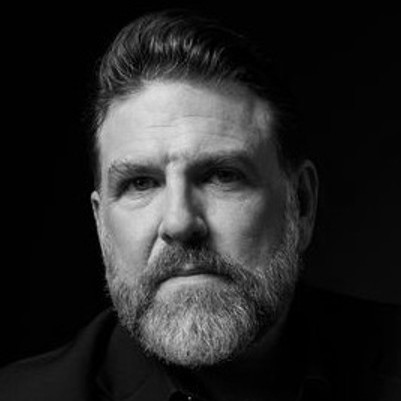 Black and white portrait of Michael Mayes, a middle-aged man with a full beard and moustache, short styled hair, and a serious expression, set against a dark background.