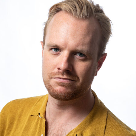 Morgan Pearse, a man with short blond hair and a light beard, wears a mustard yellow collared shirt as he looks at the camera against a plain white background.