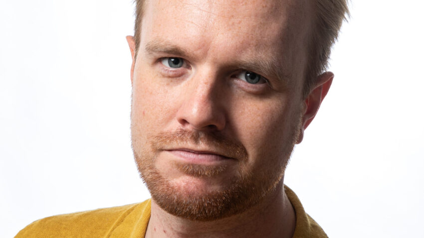 Morgan Pearse, a man with short blond hair and a light beard, wears a mustard yellow collared shirt as he looks at the camera against a plain white background.
