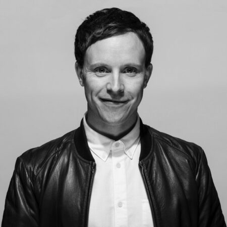 A man with short dark hair smiles slightly, wearing a white button-up shirt and a dark leather jacket, against a plain light background. The photo is in black and white.