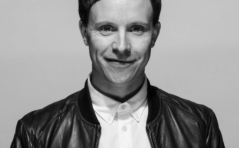 A man with short dark hair smiles slightly, wearing a white button-up shirt and a dark leather jacket, against a plain light background. The photo is in black and white.
