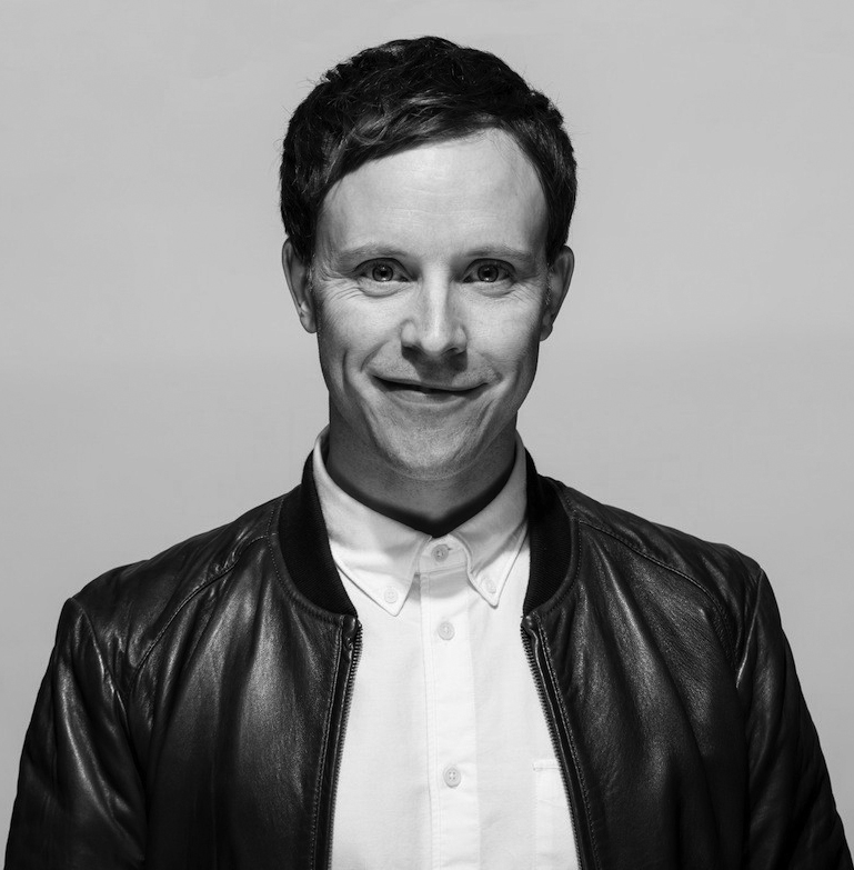 A man with short dark hair smiles slightly, wearing a white button-up shirt and a dark leather jacket, against a plain light background. The photo is in black and white.