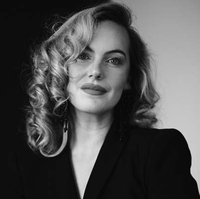 Black and white portrait of Niamh O'Sullivan with wavy, shoulder-length hair, wearing a dark blazer, looking at the camera with a slight smile and confident expression.