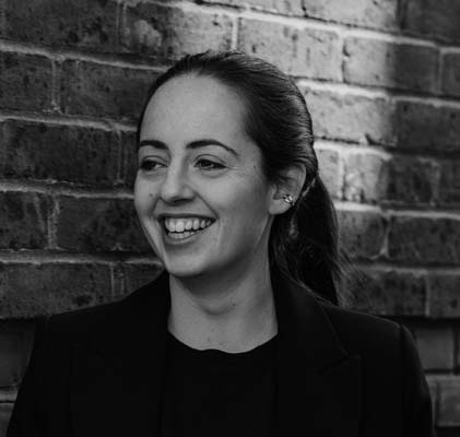 Olivia Clarke, with her dark hair tied back, smiles and looks to the side whilst standing in front of a brick wall. She is dressed in a dark top and blazer. The image is in black and white.