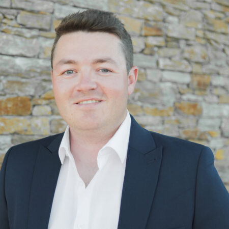 Osian Wyn Bowen, dressed in a dark suit and white shirt, stands smiling in front of a stone wall.