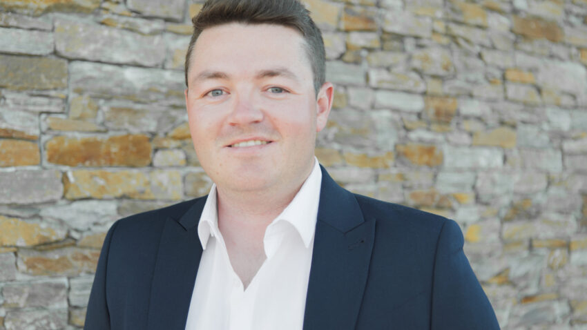 Osian Wyn Bowen, dressed in a dark suit and white shirt, stands smiling in front of a stone wall.