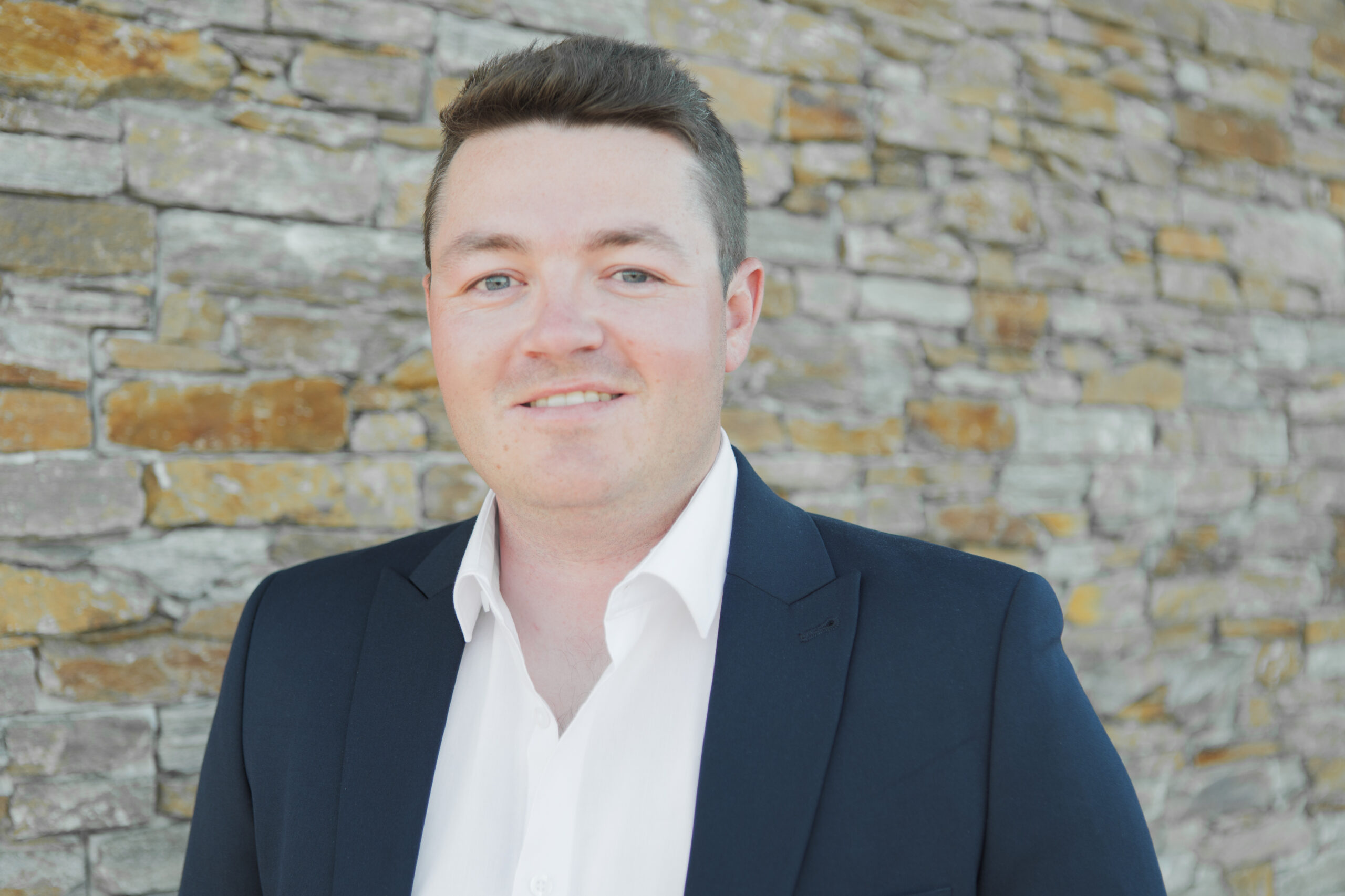 Osian Wyn Bowen, dressed in a dark suit and white shirt, stands smiling in front of a stone wall.