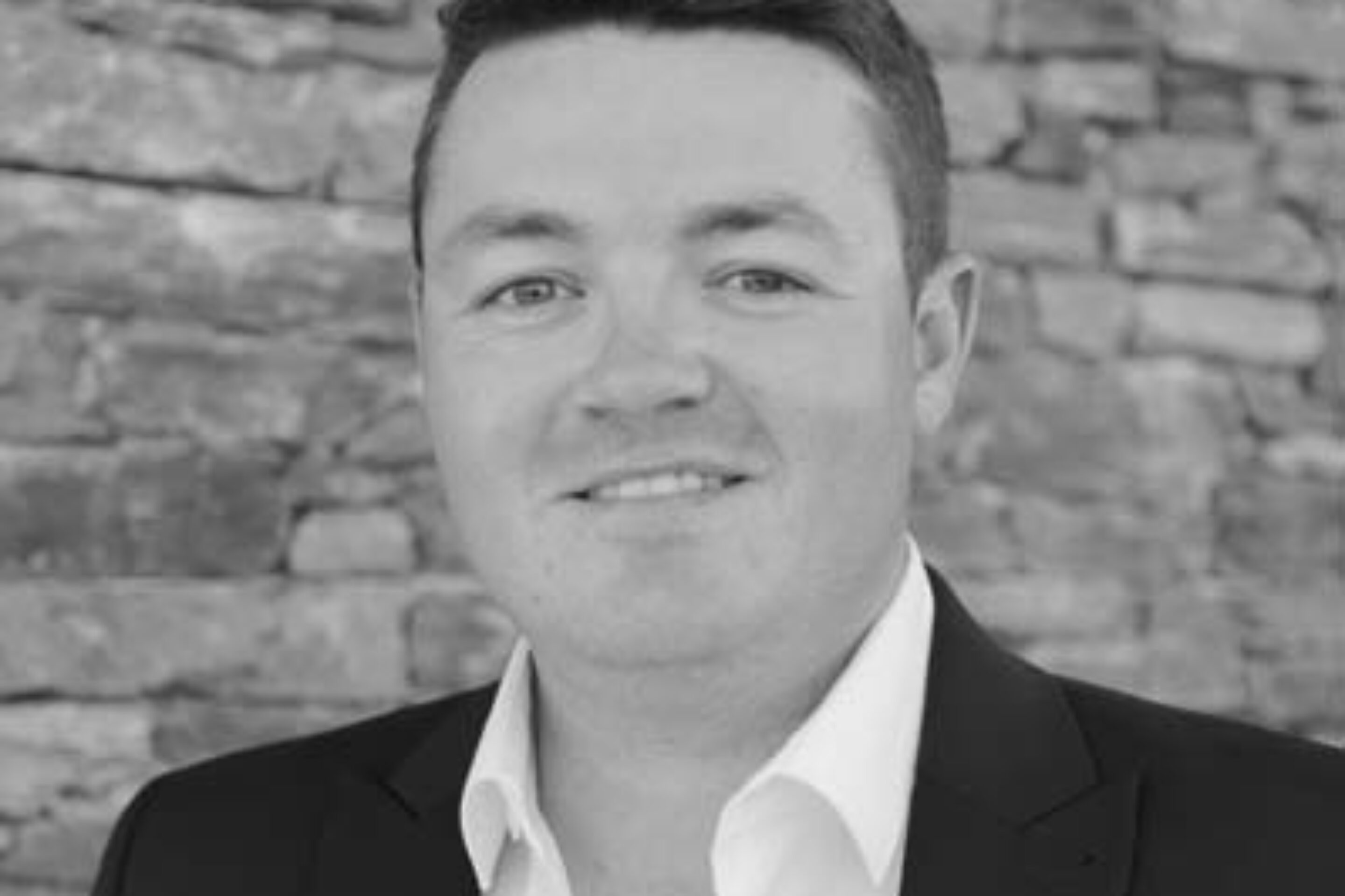 Osian Wyn Bowen, wearing a dark suit jacket and white shirt, stands in front of a stone wall and smiles slightly. The photo is in black and white.