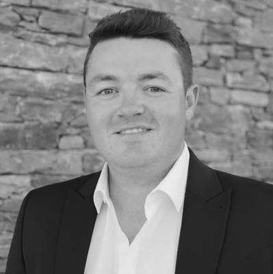 Osian Wyn Bowen, wearing a dark suit jacket and white shirt, stands in front of a stone wall and smiles slightly. The photo is in black and white.