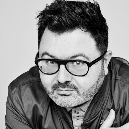 A man with short dark hair, a beard, and glasses—resembling Christian Curnyn—looks directly at the camera. He is wearing a jacket over a button-up shirt and posing against a plain background in this black and white photo.