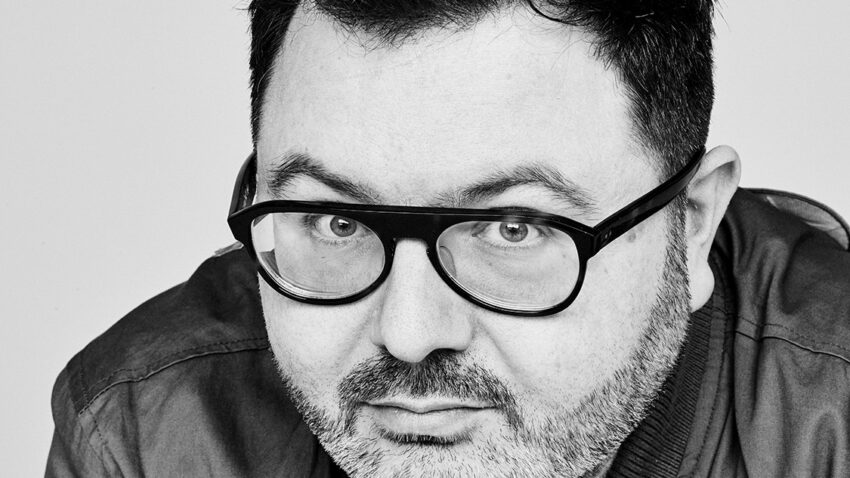 A man with short dark hair, a beard, and glasses—resembling Christian Curnyn—looks directly at the camera. He is wearing a jacket over a button-up shirt and posing against a plain background in this black and white photo.