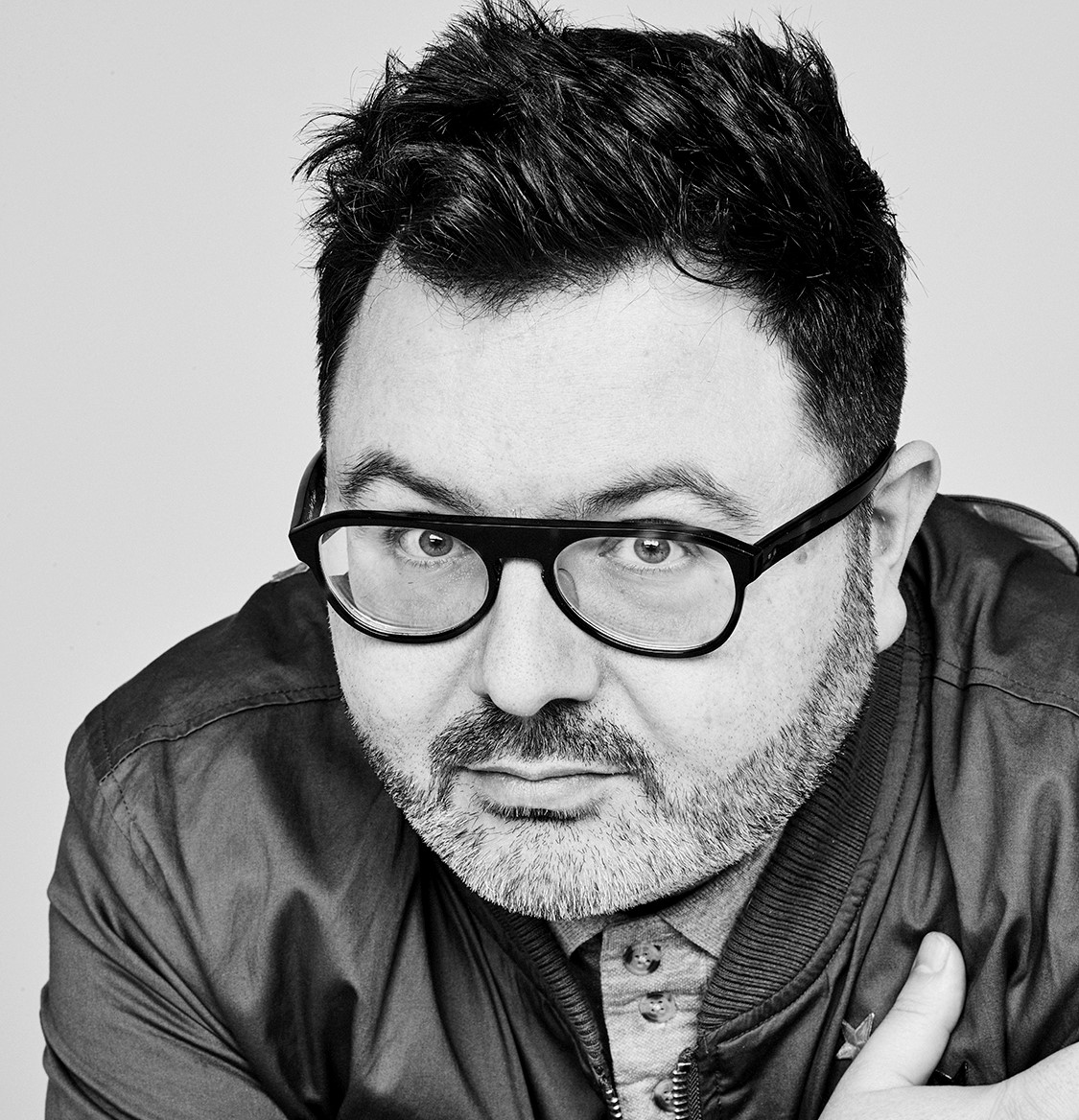 A man with short dark hair, a beard, and glasses—resembling Christian Curnyn—looks directly at the camera. He is wearing a jacket over a button-up shirt and posing against a plain background in this black and white photo.