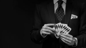 A person in a suit and tie holds a fanned-out pack of playing cards, with another card visible in their breast pocket. The black and white image echoes the drama of opera, focusing on their torso and hands.