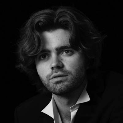 Patrick Alexander Keefe, a young man with wavy hair and a light beard, wears a suit jacket and open-collared shirt as he looks slightly upward. The black-and-white photo features a dark background, highlighting his thoughtful presence.