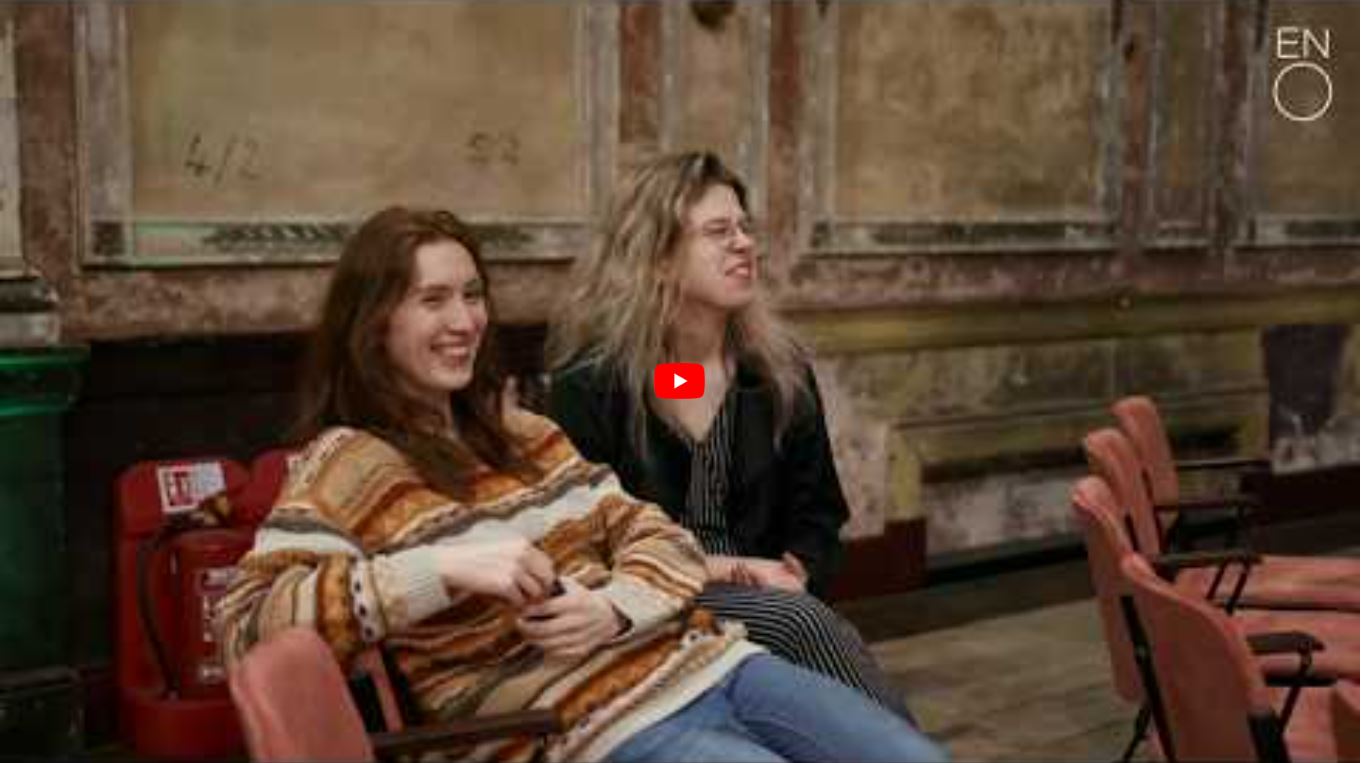 Two women sit on theatre chairs, smiling and laughing. One wears a striped jumper, the other a black jacket. The worn, textured wall behind them resembles the backdrop for an introduction to folk tales like Paul Bunyan’s.