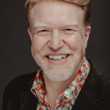 A smiling middle-aged man, resembling Paul Sheehan, with light skin, short reddish hair, and a beard, wearing a dark jacket over a colourful floral-patterned shirt, poses in front of a dark background.
