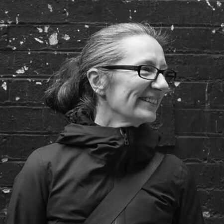 A person with glasses and long hair tied back smiles whilst looking to the side, standing against a weathered brick wall. The black and white photo.