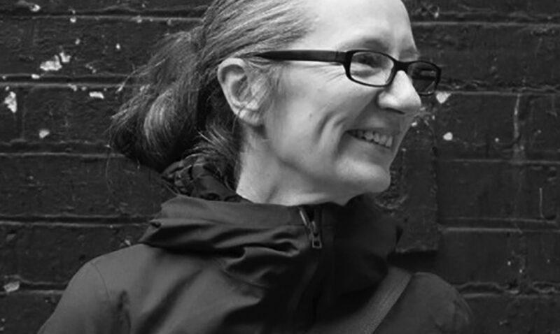 A person with glasses and long hair tied back smiles whilst looking to the side, standing against a weathered brick wall. The black and white photo.