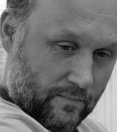 A man with a beard and short hair looks down with a thoughtful, serious expression. This black and white close-up portrait captures Peter Relton’s face in striking detail.