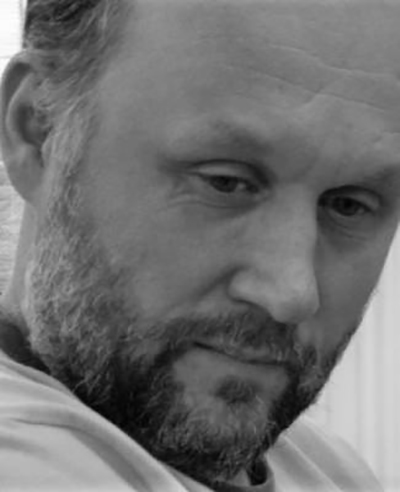 A man with a beard and short hair looks down with a thoughtful, serious expression. This black and white close-up portrait captures Peter Relton’s face in striking detail.