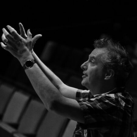 A man in a checked shirt, reminiscent of theatre director Phelim McDermott, gestures upward with both hands, palms open, whilst seated in an empty theatre. The black-and-white image captures his expressive pose and focused expression.