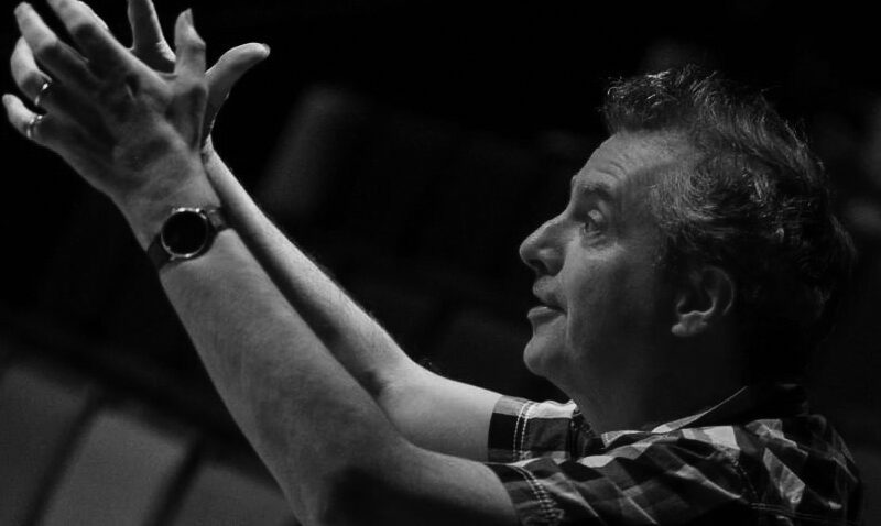 A man in a checked shirt, reminiscent of theatre director Phelim McDermott, gestures upward with both hands, palms open, whilst seated in an empty theatre. The black-and-white image captures his expressive pose and focused expression.