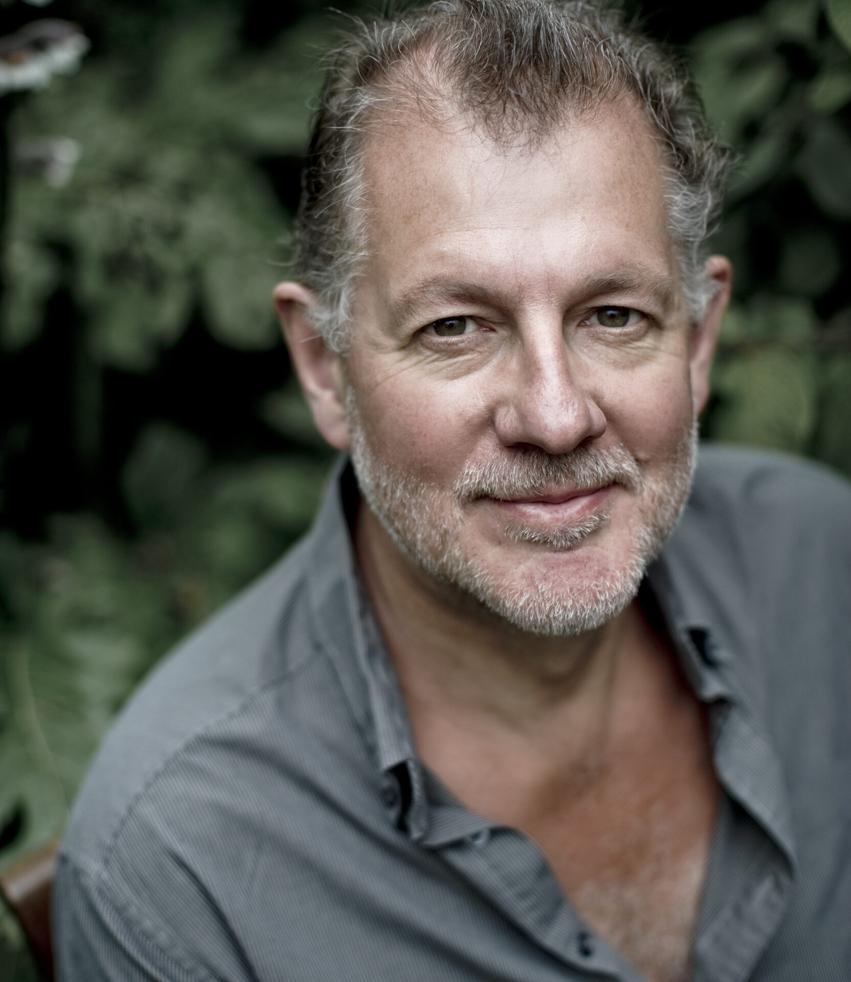 Robert Hayward, a middle-aged man with short, greying hair and a beard, smiles warmly in a partially unbuttoned grey shirt as he stands surrounded by lush, green foliage.