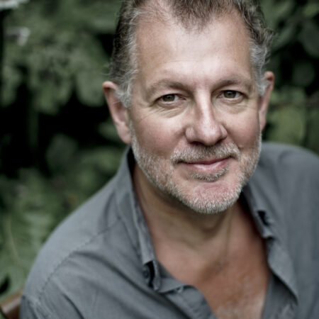 Robert Hayward, a middle-aged man with short, greying hair and a beard, smiles warmly in a partially unbuttoned grey shirt as he stands surrounded by lush, green foliage.