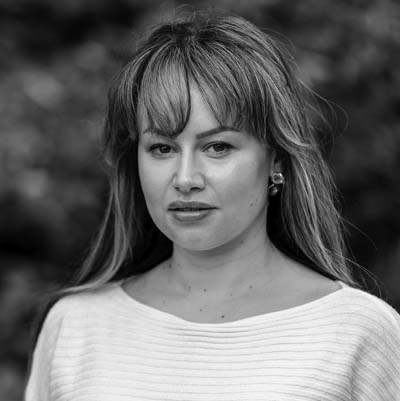 Black and white portrait of Siân Griffiths, a woman with long hair and fringe, wearing a light-coloured, textured top. She gazes directly at the camera with a neutral expression against a blurred outdoor background.