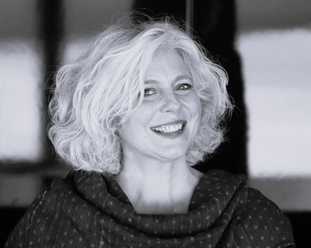 Sophie Daneman, with wavy, shoulder-length grey hair, smiles at the camera. She is wearing a dark, textured shawl. The background is blurred and appears to be indoors. The photo is in black and white.