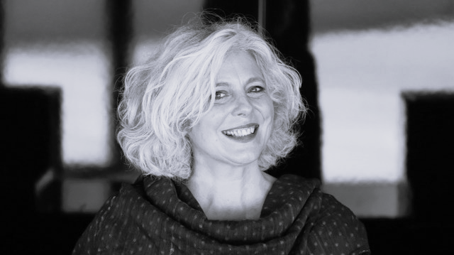 Sophie Daneman, with wavy, shoulder-length grey hair, smiles at the camera. She is wearing a dark, textured shawl. The background is blurred and appears to be indoors. The photo is in black and white.