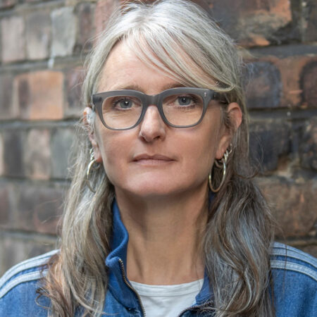 Soutra Gilmour, with long grey hair and glasses, stands in front of a brick wall wearing a blue jacket and hoop earrings, looking directly at the camera with a neutral expression.
