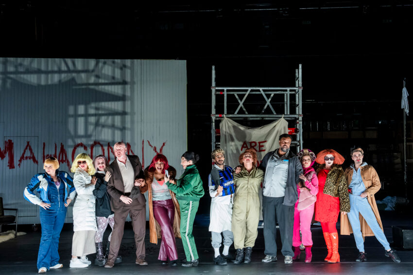 A diverse group of performers in colourful, eclectic costumes pose together on stage before a set featuring scaffolding and a painted sign that reads MAHAGONNY, evoking the dynamic energy of the City of Mahagonny in this lively theatre production.